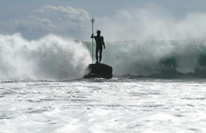 An awe-inspiring sculpture of Neptune, or Poseidon, stands firmly on a rock in Gran Canaria, Spain, as a massive wave surges behind him. The god of the sea holds his iconic trident with an air of authority, while sunlight glistens on the foaming water, capturing the raw power and beauty of the ocean.