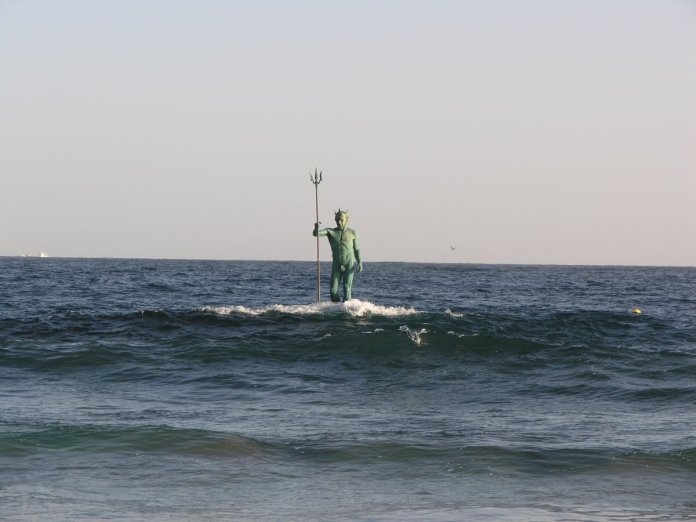 A serene view of the Neptune, or Poseidon, sculpture in Gran Canaria, Spain, standing tall and calm amidst gentle ocean waves. The sea god holds his trident, with the horizon stretching into the distance under a soft, pale sky, creating a peaceful and majestic scene.