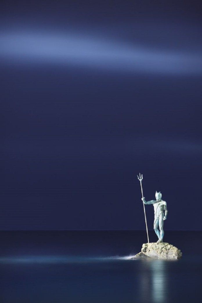 A mesmerizing nighttime view of the Neptune, or Poseidon, sculpture in Gran Canaria, Spain, standing on a rock illuminated against a deep blue sky. The calm ocean reflects the faint glow around the sea god, who holds his trident with a commanding yet serene presence, evoking the mystique of mythology under the night sky.