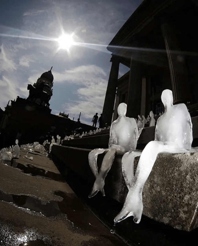 Close-up of melting ice figurines shaped like human figures sitting on the steps of Gendarmenmarkt square in Berlin, Germany. The sun shines brightly in the background, illuminating the scene and casting shadows. The sculptures, part of Nele Azevedo’s climate change art installation, symbolize the fragility of human life and the urgent need to address global warming. The historic architecture of the square and silhouettes of onlookers add depth to the striking visual message.