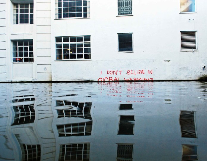 Street art by Banksy in Camden, London, showing the text 'I DON’T BELIEVE IN GLOBAL WARMING' written in red graffiti on a white building wall. The text is partially submerged in water, symbolizing rising sea levels. The reflection of the graffiti and the building is visible in the water, creating a striking visual commentary on climate change.