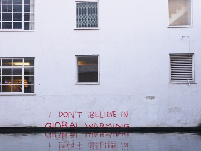 Street art by Banksy in Camden, London, featuring the text 'I DON’T BELIEVE IN GLOBAL WARMING' written in bold red graffiti on a white building wall. The text appears just above the waterline, emphasizing the reality of rising sea levels. The wall's windows and reflections in the water contribute to the powerful environmental message.
