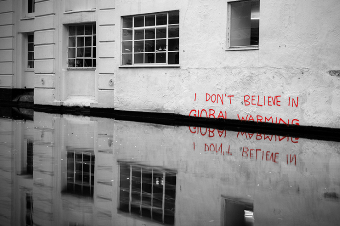 Street art by Banksy in Camden, London, featuring the text 'I DON’T BELIEVE IN GLOBAL WARMING' in bold red graffiti on a white wall. The partially submerged message reflects in the water below, emphasizing the pressing issue of climate change and rising sea levels. The black-and-white background highlights the vibrant red text, drawing attention to the artwork's stark message.