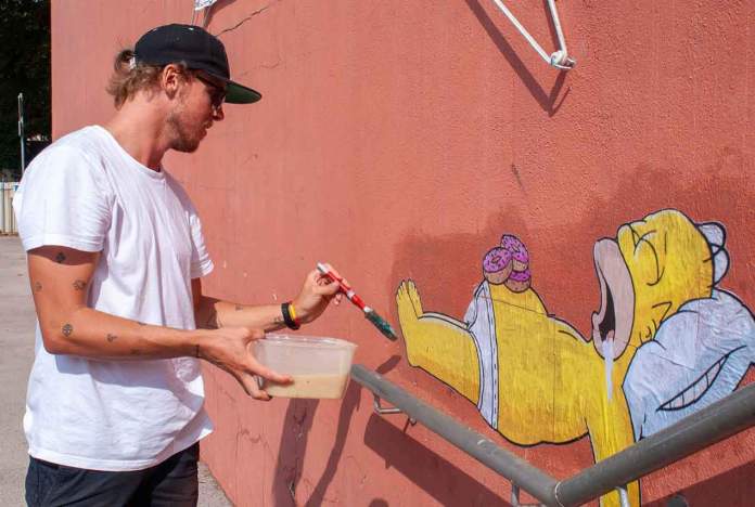 EFIX, the street artist, is seen applying his artwork of Homer Simpson lounging on a railing. He is holding a brush in one hand and a container with paste in the other, carefully attaching the piece to a red wall. Homer, depicted in his iconic lazy pose with donuts and drool, humorously integrates with the urban railing. The scene highlights EFIX’s artistic process and his playful, eco-friendly approach to street art.