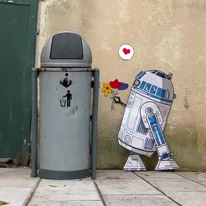 A street art piece by EFIX showing R2-D2 from Star Wars offering a bouquet of colorful flowers to a gray trash can. A red heart speech bubble floats above R2-D2, adding a touch of humor and warmth. The art is placed against a worn beige wall, blending urban elements with pop culture in a playful way.