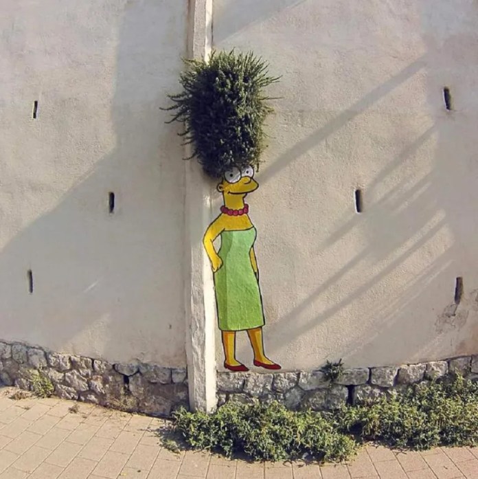 A street art piece by EFIX featuring Marge Simpson from The Simpsons, painted on a wall with her iconic tall blue hair creatively formed by a real bush growing above her. Marge is depicted in her signature green dress and red necklace, seamlessly integrating with the natural and urban elements of the setting. The artwork humorously blends the living plant with the character, bringing a playful and unexpected charm to the environment.