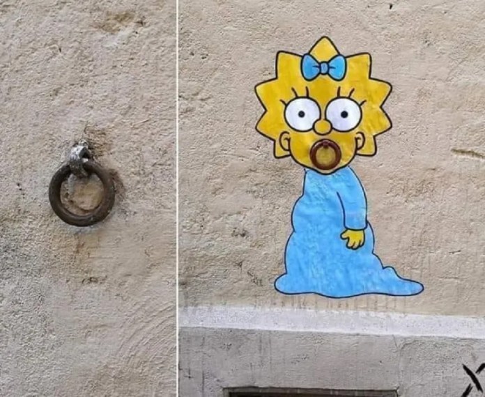 A street art piece by EFIX featuring Maggie Simpson from The Simpsons, painted on a wall with her pacifier cleverly integrated into a metal ring attached to the surface. Maggie is depicted in her signature blue outfit and bow, humorously interacting with the urban element. The artwork demonstrates EFIX’s playful ability to merge cartoon characters with everyday street objects.
