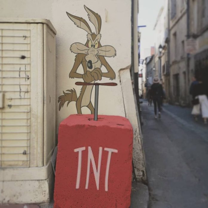 A street art creation by EFIX featuring Wile E. Coyote from Looney Tunes, painted on a wall. The piece captures Wile E. Coyote's signature mischievous expression, blending humor with the surrounding street environment. The background includes a narrow alley with pedestrians, emphasizing the playful interaction between the art and its urban setting.