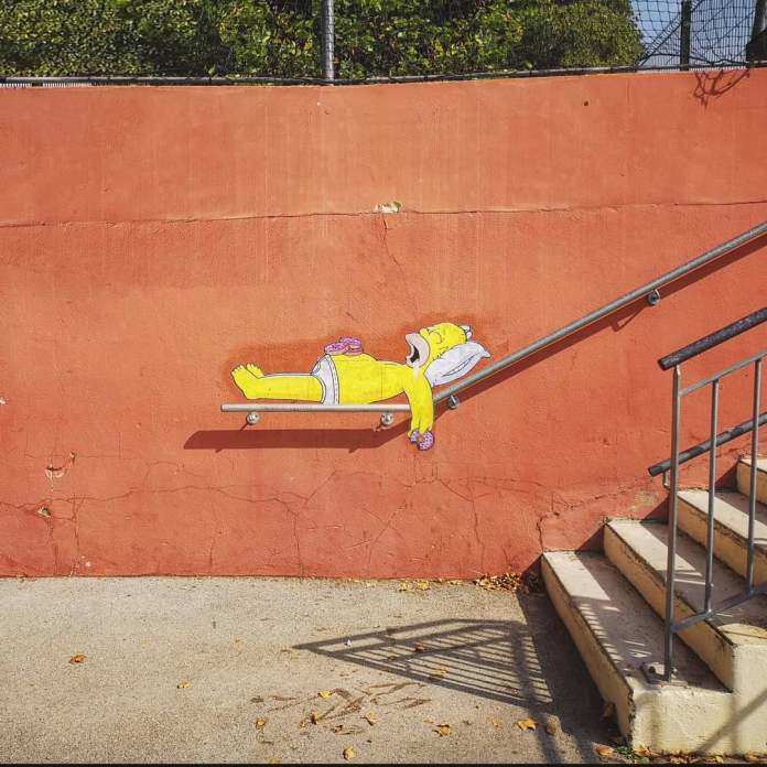 A street art piece by EFIX featuring Homer Simpson from The Simpsons, creatively integrated into a railing on a staircase. Homer is depicted lounging in his underwear on the inclined railing, using it as a makeshift bed, complete with a pillow and slippers. The artwork blends humor with the environment, transforming the urban feature into a playful and unexpected scene. The background consists of a red wall and nearby stairs, adding depth to the composition.