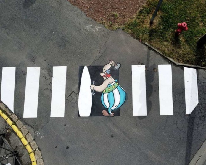 A playful street art piece by Oakoak featuring Obelix, the beloved character from the Asterix comics, creatively integrated into a pedestrian crossing. The white stripes of the crosswalk are transformed into the menhir Obelix carries on his shoulder, blending urban infrastructure with whimsical artistry. This artwork highlights Oakoak's signature style of reimagining urban elements into engaging and humorous scenes. The setting includes surrounding asphalt, a yellow curb, and a patch of grass with a red fire hydrant in the background.