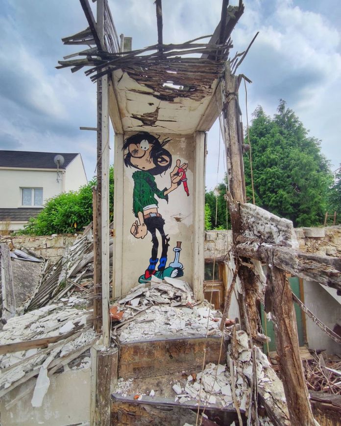A striking street art piece by Oakoak featuring Gaston Lagaffe, a classic comic character, painted on the interior wall of a dilapidated, partially destroyed building. Gaston is depicted holding a red marker, with his signature disheveled appearance, adding a humorous and creative touch to the otherwise crumbling environment. Surrounding the artwork are piles of rubble, broken wood, and remnants of the building's structure, with green foliage and neighboring houses visible in the background. This piece exemplifies Oakoak's ability to inject playfulness into abandoned urban spaces.