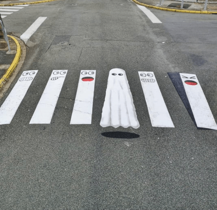 A whimsical street art piece by Oakoak transforming a pedestrian crosswalk into a playful scene featuring cartoon faces and a ghost. The white stripes of the crosswalk are painted with various expressive faces, while one stripe is creatively turned into a floating ghost, complete with a shadow beneath it for a 3D effect. The artwork adds humor and life to an ordinary urban setting, surrounded by asphalt, a yellow curb, and intersecting streets. This piece showcases Oakoak's talent for injecting fun and creativity into everyday public spaces.
