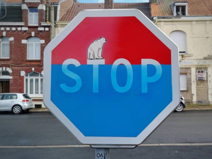 A thought-provoking street art piece by Oakoak that transforms a stop sign into a commentary on climate change. The sign is painted with a red and blue gradient, suggesting rising water levels, and features a polar bear stranded on a small ice block above the word "STOP." The artwork cleverly integrates the message into the urban landscape, set against a background of brick buildings and parked cars. This piece highlights Oakoak's ability to blend environmental themes with urban street art in a visually impactful way.
