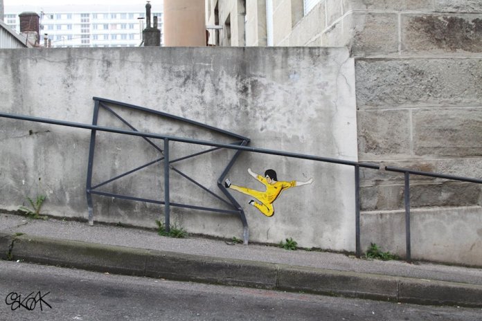 A creative street art piece by Oakoak depicting Bruce Lee mid-action, dressed in his iconic yellow jumpsuit, kicking through a bent metal railing. The artwork cleverly uses the damaged railing to enhance the dynamic motion of the martial arts move, making it appear as though Bruce Lee has just broken it with his powerful kick. Set against a concrete wall in an urban environment, this piece exemplifies Oakoak's talent for transforming everyday urban structures into humorous and interactive scenes.