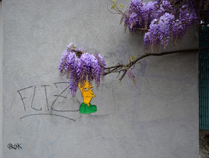 A witty street art piece by Oakoak featuring a character resembling Sideshow Bob from The Simpsons with wisteria flowers cleverly placed as his hair. The painted face is set against a plain gray wall, with the vibrant purple wisteria blooms extending from a nearby branch to complete the humorous depiction. The artwork seamlessly blends natural elements with urban art, showcasing Oakoak's creativity in integrating surroundings into his playful designs.