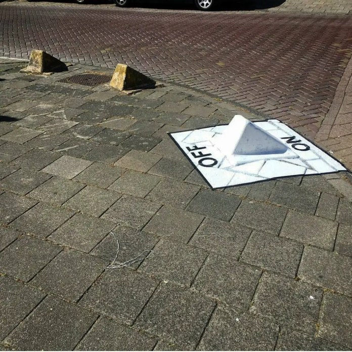 Street artwork by Oakoak depicting a large "ON/OFF" switch painted on the pavement, with a raised white element creating the illusion of a functional button.