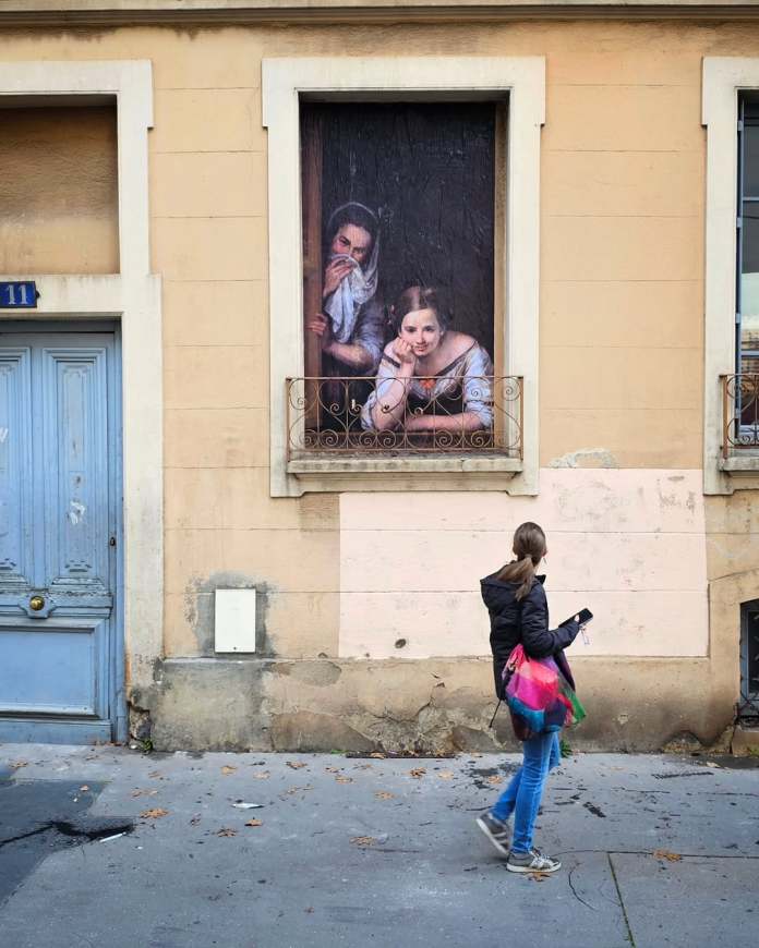A street art piece by Oakoak incorporating a real balcony railing, with a painted figure extending from it, creating the illusion of a person standing casually on the balcony.