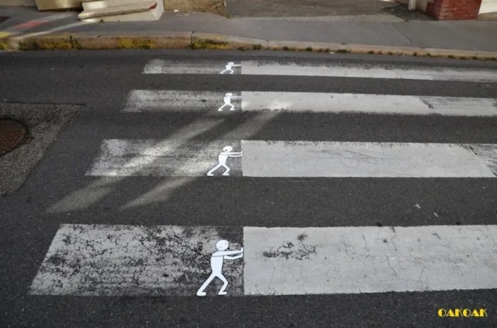 A street art intervention by Oakoak on a pedestrian crosswalk, depicting small white stick figures seemingly pushing the worn-out stripes of the crossing.