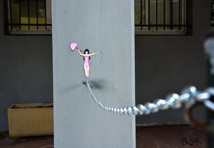 A street art piece by Oakoak depicting a small illustrated woman in a pink dress, balancing on a real metal chain as if performing a tightrope act, holding a tiny umbrella.