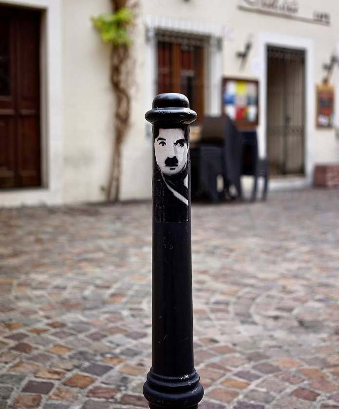 A street art intervention by Oakoak featuring a black bollard painted with the face and hat of Charlie Chaplin, playfully turning an ordinary street post into an artistic tribute.