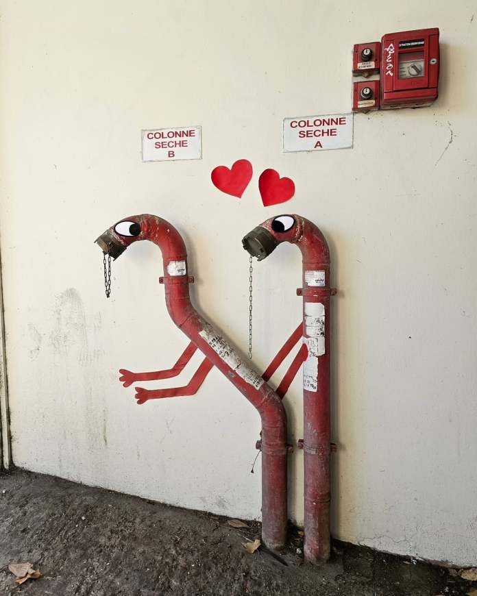 A street art piece by Oakoak featuring a small painted sign reading "Very Hot" placed near a burn mark on a wall, making it appear as if the scorch damage is part of an official cautionary notice.