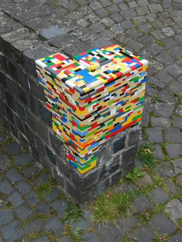 "A close-up of a street art installation by Jan Vormann's Dispatchwork project, where a corner of a weathered stone wall is creatively patched with vibrant LEGO bricks. The colorful LEGO repair contrasts beautifully with the dark, aged stones and cobblestone pavement below, blending playful design with the textures of urban decay.