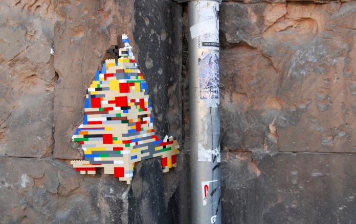 A whimsical LEGO installation from Jan Vormann's Dispatchwork project, shaped like a colorful face silhouette, seamlessly integrated into a crack in a weathered stone wall. The vibrant LEGO bricks, featuring red, blue, yellow, and green, stand out against the muted tones of the natural stone. A nearby metal pole covered in stickers and graffiti adds an urban touch to the scene.