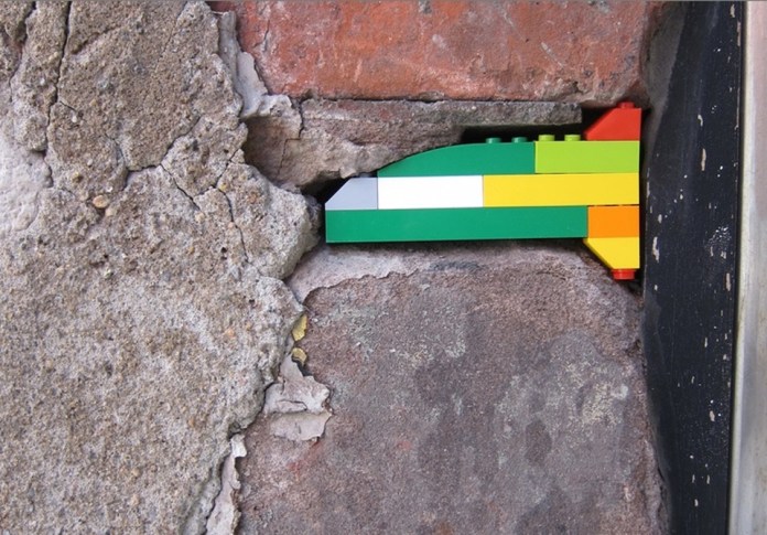 A small and subtle LEGO repair from Jan Vormann's Dispatchwork project, filling a crack in an old, weathered stone wall. The LEGO bricks, arranged in green, yellow, white, and orange, form a clean and colorful line that contrasts with the rugged texture of the surrounding wall. The repair blends functionality with artistic charm.