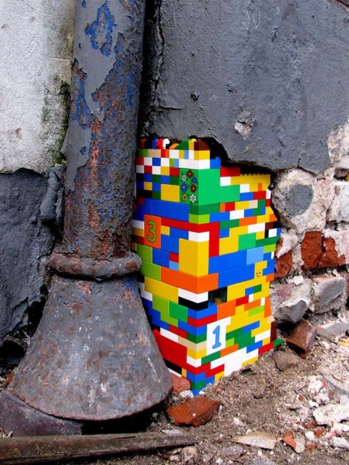 A vibrant LEGO repair from Jan Vormann's Dispatchwork project, filling a corner of a crumbling wall with multicolored bricks. The repair includes numbered LEGO pieces, such as '3' and '1,' and features small decorative flower prints. It contrasts sharply with the aged, rusted metal pipe and the peeling gray paint of the surrounding wall, adding a playful touch to the urban decay.