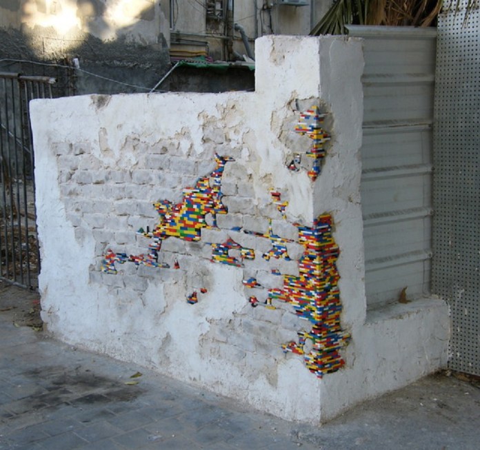 A weathered white plaster wall repaired with colorful LEGO bricks as part of Jan Vormann's Dispatchwork project. The LEGO pieces fill cracks and missing bricks throughout the structure, creating a striking mosaic of red, blue, yellow, green, and other colors. The playful repairs contrast with the aged and deteriorating surface of the wall, bringing a vibrant artistic element to the urban environment.