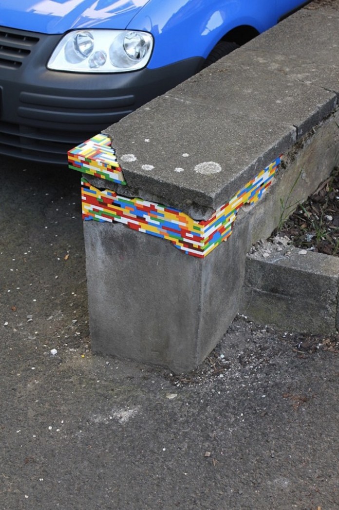 A concrete pillar repaired with colorful LEGO bricks as part of Jan Vormann's Dispatchwork project. The vibrant LEGO pieces fill the missing section at the top, standing out against the dull gray cement and blending creativity with functionality. A blue car is parked nearby, adding to the urban setting of this playful intervention.