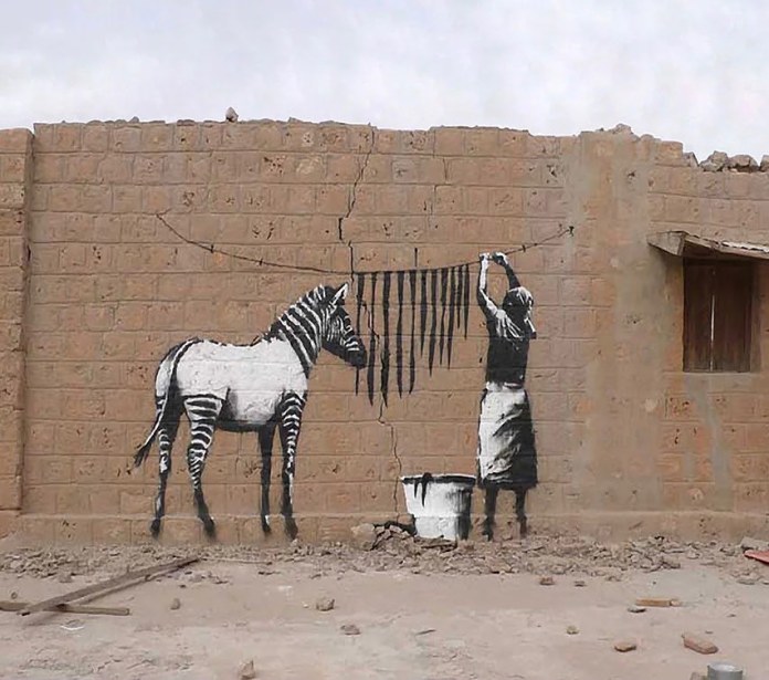 A striking piece of street art by Banksy on a cracked brick wall in a rural setting. The artwork features a zebra and a woman cleverly interacting with their environment. The woman hangs striped laundry on a clothesline, aligning perfectly to form the zebra's body stripes. A bucket of paint sits on the ground below. This iconic Banksy piece blends humor and creativity, seamlessly integrating the wall's texture into its storytelling.