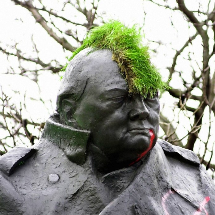 A statue of Winston Churchill modified with humorous and rebellious touches. The statue, crafted from dark material, features a bright green patch of grass styled as a mohawk on the head, adding a punk aesthetic. Additionally, red paint drips down from the mouth, resembling blood. The background showcases leafless tree branches, emphasizing the outdoor setting. The alterations create a provocative and satirical commentary, blending historical reverence with modern rebellious art.