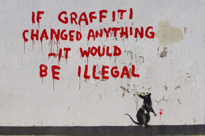 A Banksy mural featuring the phrase "If graffiti changed anything - it would be illegal" painted in bold red letters on a white, cracked wall. Below the text, a black stencil of a rat appears, holding a paintbrush with red paint dripping from it, as though the rat created the graffiti. The image critiques the societal view of graffiti and its perceived impact on change.