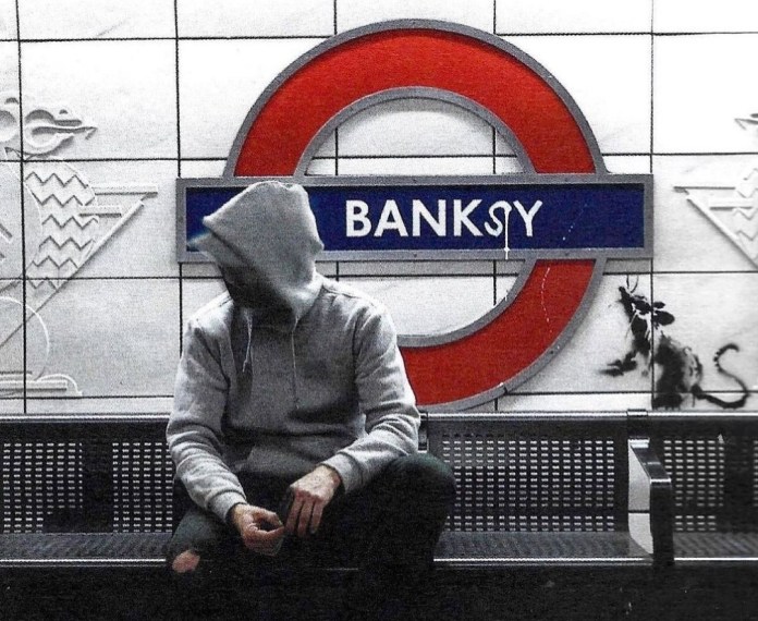 "A person in a gray hoodie sits on a bench at what appears to be a London Underground station, with the iconic red-and-blue roundel logo altered to read 'BANKSY.' Behind them, a black stencil of a rat, characteristic of Banksy's graffiti, is visible on the tiled wall.