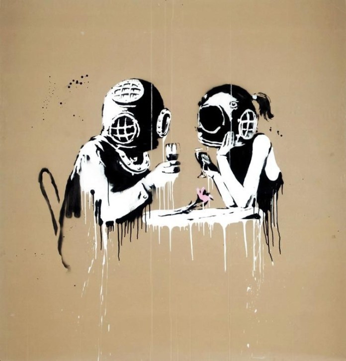 Street art piece depicting two people wearing vintage diving helmets, sitting across from each other at a table, each holding a glass. The artwork is characterized by drips of paint, giving it a raw, unfinished look, while a small pink flower adds a touch of color and tenderness to the scene. The piece evokes themes of isolation and connection, possibly hinting at barriers in communication or intimacy.
