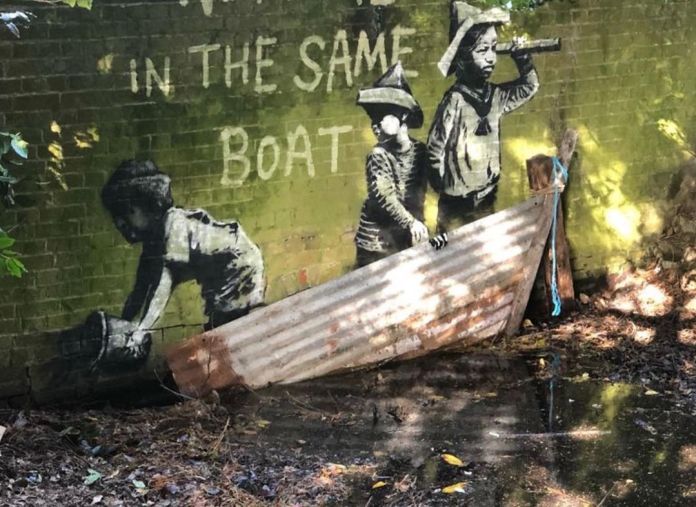 A closer view of Banksy's mural titled "We're All in the Same Boat" on a mossy brick wall in Oulton Broad, England. The artwork depicts three children in monochrome: one crouched and reaching into the water, one standing and holding a telescope, and one looking ahead, all wearing paper hats. In front of the mural, a rusted corrugated metal piece, resembling a makeshift boat, is propped against the wall, adding a three-dimensional element to the scene. The text "WE'RE ALL IN THE SAME BOAT" is painted in white above the figures.