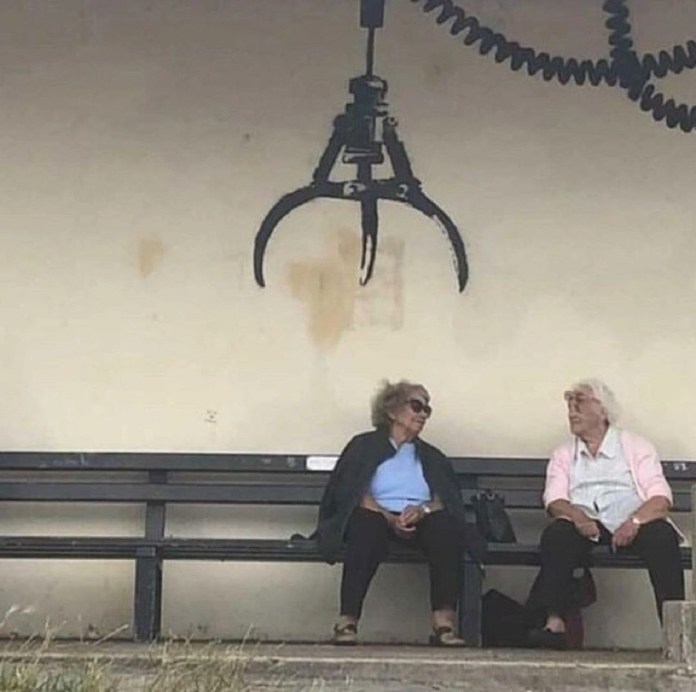 A Banksy mural titled "Arcade Claw Machine" on a public wall in Gorleston, England. The artwork depicts a black arcade-style claw machine hanging from a coiled cord, painted above a bench. Two elderly women are seated on the bench, engaged in conversation, adding a whimsical juxtaposition between the playful, mechanical claw and the calm, everyday scene. The mural cleverly integrates the real-world bench and setting into the artwork.