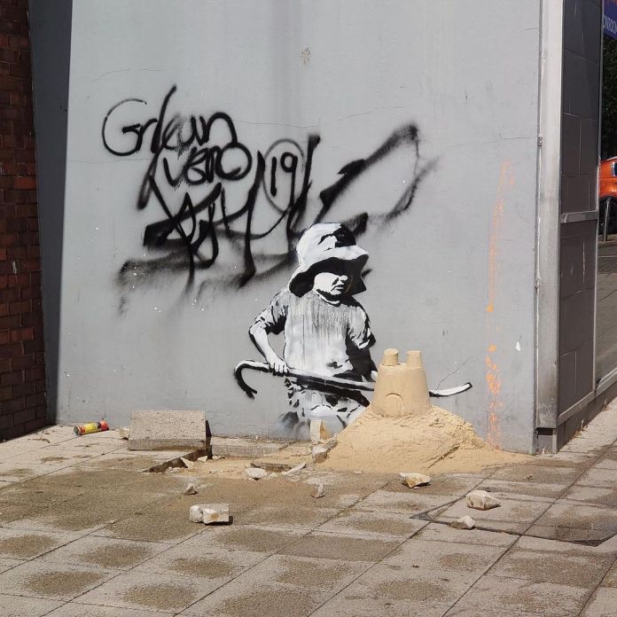 A Banksy mural titled "Child with Sandcastle" on a street corner in Lowestoft, England. The artwork features a monochrome depiction of a young child wearing a floppy hat, holding a crowbar, and standing beside a realistic sandcastle. The sandcastle is sculpted from actual sand and sits on a damaged paving slab, blending the mural with its physical surroundings. The wall behind the child features black graffiti tags, contrasting with the intricate and thoughtful Banksy piece.