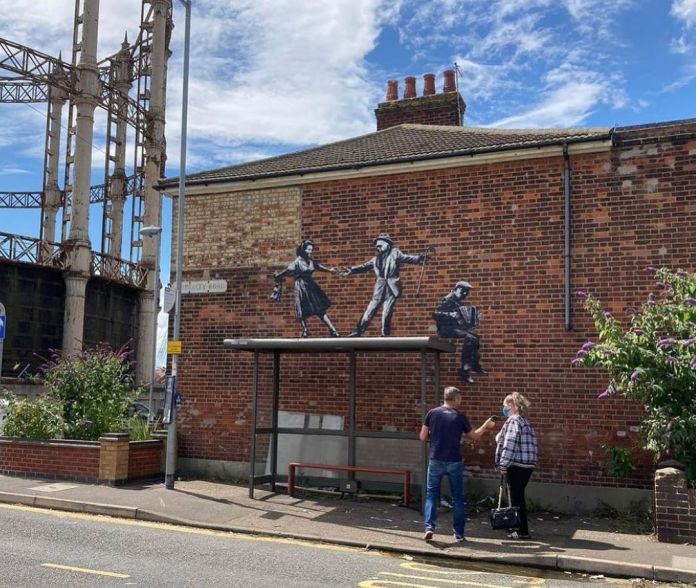 A Banksy mural titled "Dancing Couple and Accordion Player" on a brick wall in Great Yarmouth, England. The artwork depicts a monochrome couple dressed in vintage attire dancing gracefully on top of a real bus stop, accompanied by a seated accordion player painted on the wall. The scene is located near industrial gas holders and greenery, blending urban and natural elements. Two people stand in front of the bus stop, adding a sense of scale and interaction to the artwork.