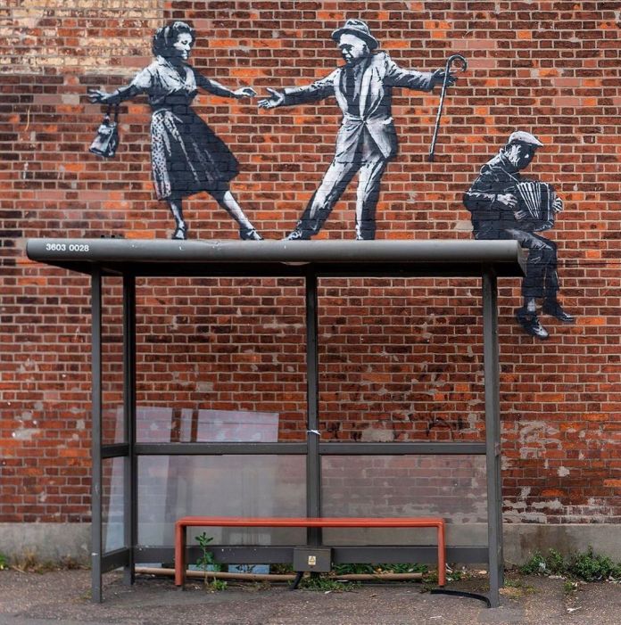 A closer view of Banksy's mural titled "Dancing Couple and Accordion Player" on a red brick wall in Great Yarmouth, England. The artwork features a monochrome couple in vintage clothing dancing on top of a real bus stop shelter, with the man holding a cane and the woman swinging a handbag. To the right, a painted accordion player sits at the edge of the bus stop roof, creating a lively and dynamic composition. The details of the artwork stand out against the textured brick wall, with the bus stop structure seamlessly incorporated into the scene.