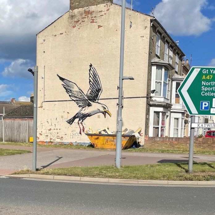 A Banksy mural featuring a giant seagull swooping down, painted on the side of a building in Lowestoft, England. The seagull appears to be scavenging for food from a real yellow skip placed in front of the mural, which is filled with debris that resembles chips. The artwork cleverly integrates the skip into the scene, creating a playful illusion. The surrounding area includes a road with a green directional sign, a lamppost, and residential buildings under a bright blue sky.