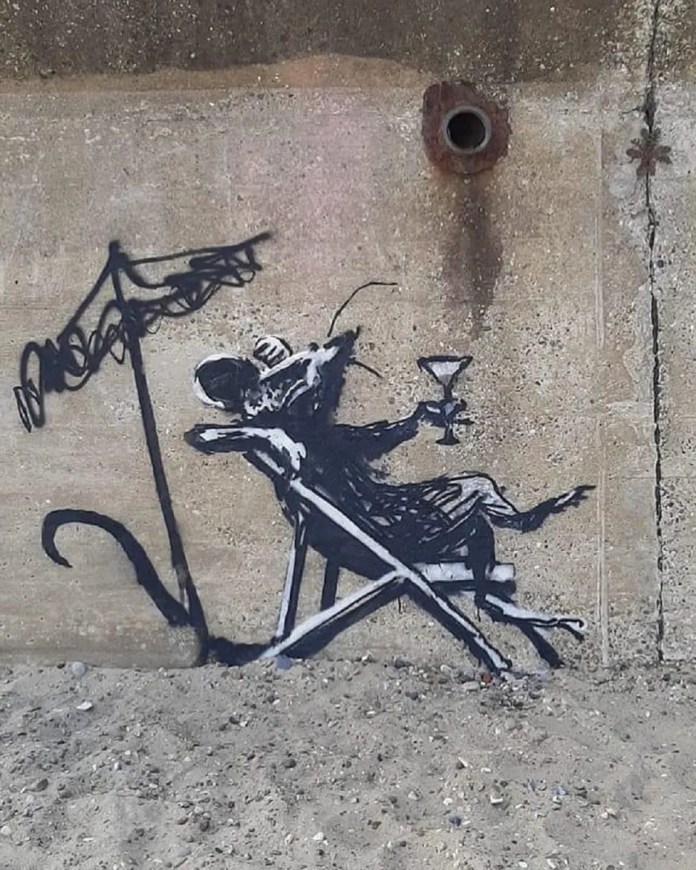 A Banksy mural titled "Rat in a Deckchair" on a concrete wall at Lowestoft North Beach, England. The artwork depicts a black and white rat lounging in a deckchair under a scribbled beach umbrella while holding a martini glass, evoking a relaxed vacation vibe. The setting includes sand at the base of the wall, blending the mural seamlessly into the seaside environment. The wall features natural cracks and a rusted pipe, adding to the raw and unpolished aesthetic.