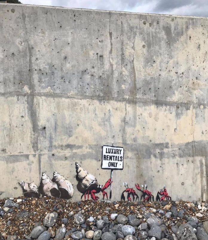 A Banksy mural titled "Luxury Rentals Only" on a concrete seawall in Cromer, England. The artwork features a group of hermit crabs, each occupying painted seashells, with one crab holding a sign that reads "Luxury Rentals Only." The mural is located at the base of the wall, surrounded by a mix of pebbles and small rocks, blending naturally into the coastal setting. The playful commentary on housing and exclusivity contrasts with the rugged and industrial appearance of the seawall.