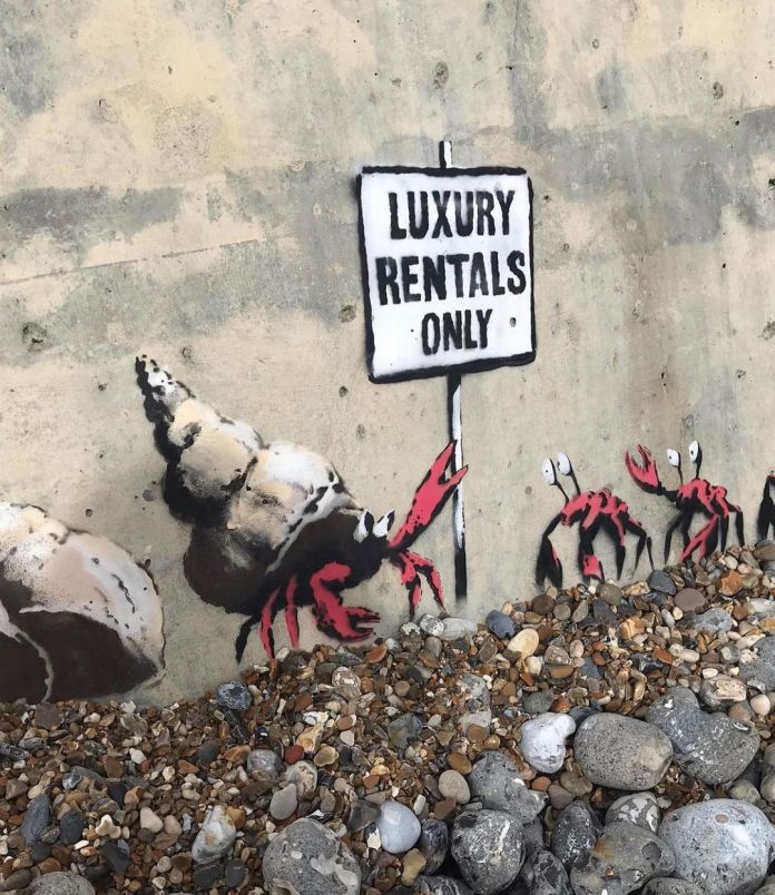 A close-up of Banksy's mural titled "Luxury Rentals Only" on a concrete seawall in Cromer, England. The detailed view highlights a hermit crab with bright red claws emerging from a painted seashell, holding a sign that reads "Luxury Rentals Only." Additional crabs with expressive eyes are visible to the right, surrounded by a pile of pebbles and stones that blend naturally into the coastal environment. The mural's humorous commentary on housing exclusivity contrasts with the raw texture of the concrete wall.