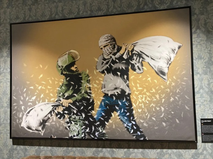 'Cushion War,' a painting by Banksy from The Walled Off Hotel exhibition in Palestine, 2017. The artwork humorously depicts a pillow fight between two figures: a soldier in military gear on the left and a civilian with a keffiyeh scarf covering his face on the right. Each figure holds a pillow mid-swing, with feathers flying through the air, symbolizing an ironic and lighthearted take on conflict in a setting typically associated with tension. The soft feathers against a warm backdrop add a surreal twist, contrasting the typical imagery of resistance with a scene of childlike play.