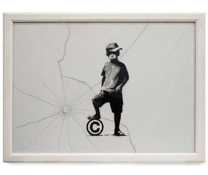 'Copyright Boy' by Banksy, created in 2012. The artwork shows a young boy standing with one foot on a large '©' symbol, symbolizing copyright, as if it's a toy ball. The glass frame around the artwork is deliberately cracked, radiating from the area near the symbol, adding a raw, rebellious element to the piece. The image critiques the commercialization and ownership of art, juxtaposing the innocence of childhood with the concept of intellectual property.