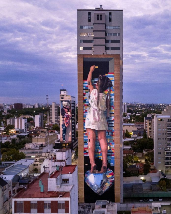 A monumental mural by Martín Ron in Banfield, Buenos Aires, Argentina. This artwork features a young girl in a floral dress standing barefoot on a reflective, heart-shaped sculpture. She reaches up with a yellow paint roller, seemingly painting a vibrant, pixelated background of multicolored blocks that extends across the tall building. The striking mural is part of an urban landscape, surrounded by city architecture under a pastel-hued evening sky. A smaller mural by Martín Ron can also be seen on a neighboring building in the background, adding depth to the scene.