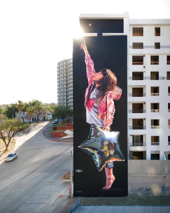 A stunning mural titled "Olivia Looks at the Sky" by Martín Ron, located in Córdoba, Argentina. The artwork depicts a young girl with long hair wearing a pink jacket and holding a shiny, reflective silver star-shaped balloon. She stands barefoot, reaching upwards with one hand as if pointing to the sky. The mural covers the side of a tall building, with sunlight dramatically peeking over the rooftop. The vibrant colors and hyper-realistic details contrast beautifully with the clean lines of the surrounding modern architecture and the open street below.