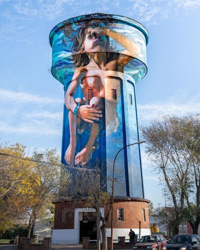 A breathtaking mural by Martín Ron on a 35-meter-high water tank in Miramar, Argentina. The artwork portrays a young boy wearing a swimsuit, captured in a serene underwater moment as she holds her nose and descends gracefully. The mural masterfully incorporates the cylindrical shape of the tank, creating the illusion of water ripples enveloping the structure. The vibrant blue tones blend harmoniously with the sky, while the realistic details make the image appear lifelike. The brick base of the tank adds a charming contrast, grounding the towering art piece in the surrounding neighborhood.
